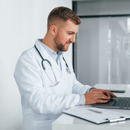 Using laptop. Professional medic in white coat is in the clinic Using laptop. Professional medic in white coat is in the clinic